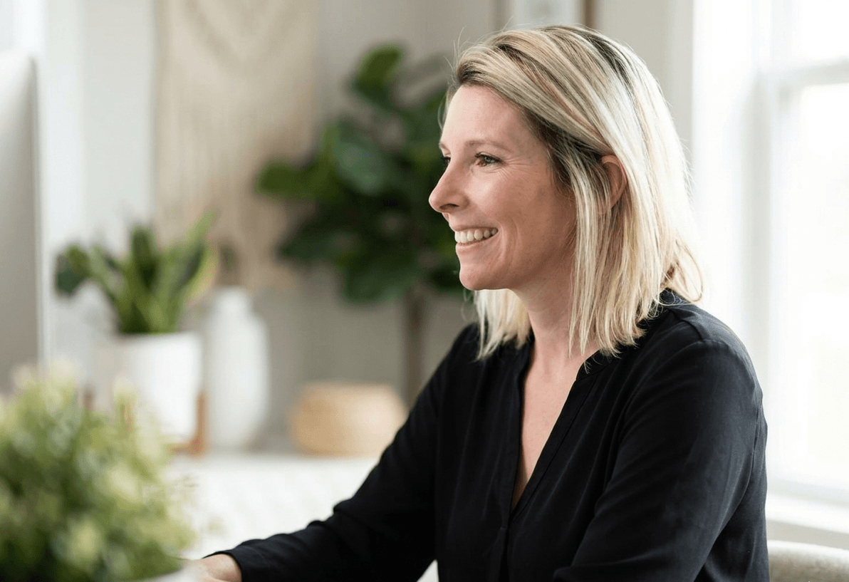 A woman with blonde shoulder-length hair wearing a black blouse and a smartwatch smiles while looking off to the side. She is seated in a bright, airy space with softly blurred green plants and neutral décor in the background.