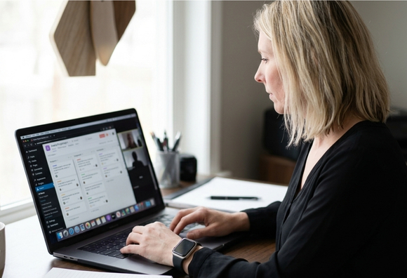 A woman with blonde shoulder-length hair wearing a black blouse and a smartwatch types on a laptop at a wood desk. The screen displays a project management interface. A notebook, pen, and cup are visible on the desk beside her, with a bright window in the background.