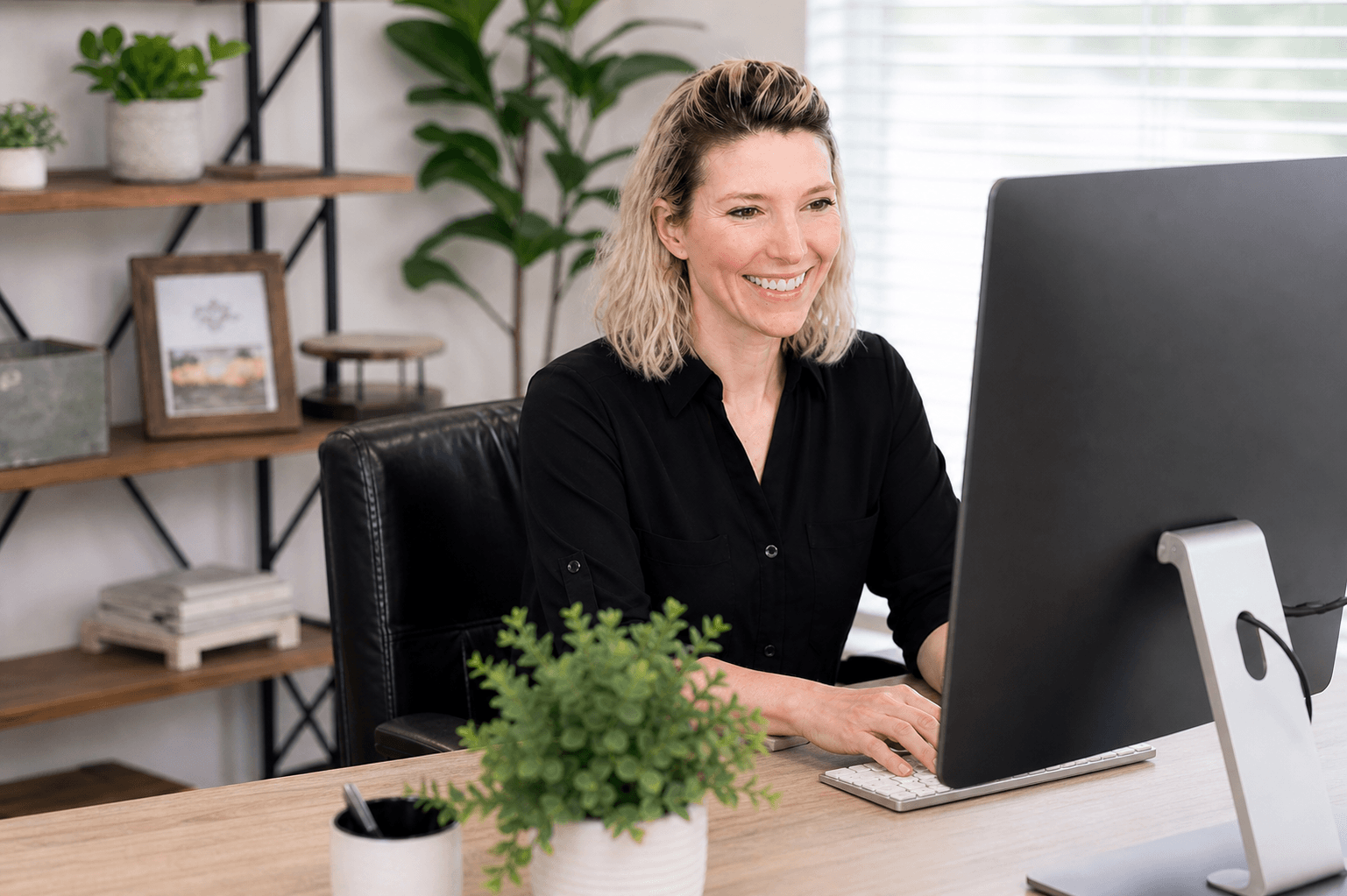 A smiling woman with blonde hair sits at a wood desk working at a desktop computer in a bright, modern home office. She is wearing a black button-up shirt and seated in a black leather chair. The background features open shelving with books, decorative items, and green plants. A small potted plant sits in the foreground on the desk.