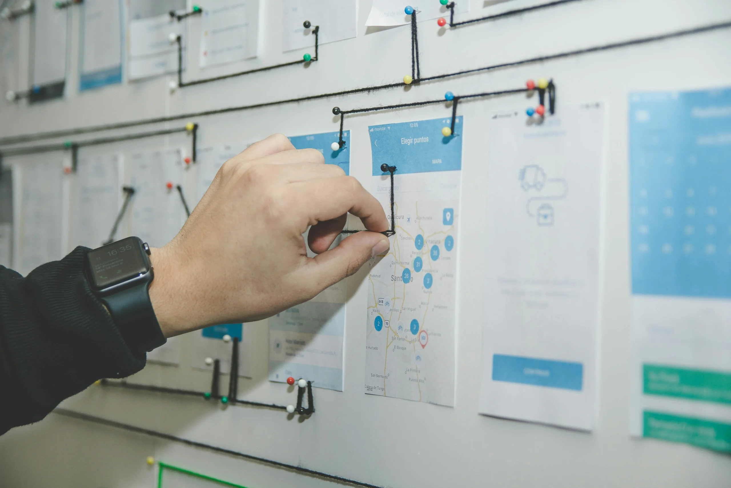 A hand wearing a smartwatch pins printed documents and wireframes to a project planning board connected with black string, representing the structured operations and project management support a PMP-certified virtual assistant provides to small businesses.