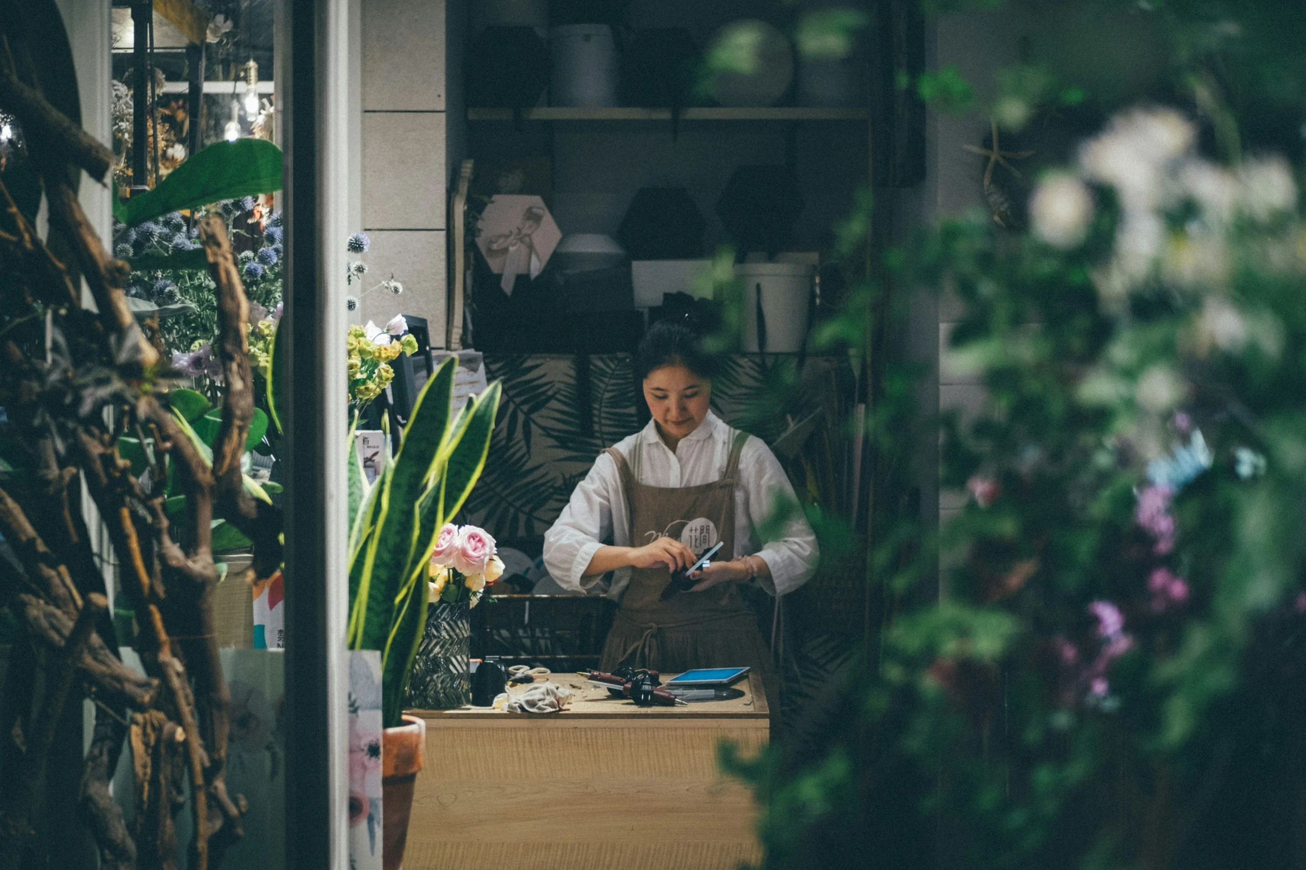A small business owner woman wearing a white blouse and brown apron stands behind the counter of a small floral and plant shop, looking down at a mobile device in her hands. She is framed by lush green plants, branches, and flowers in the foreground, giving the image a layered, intimate feel.