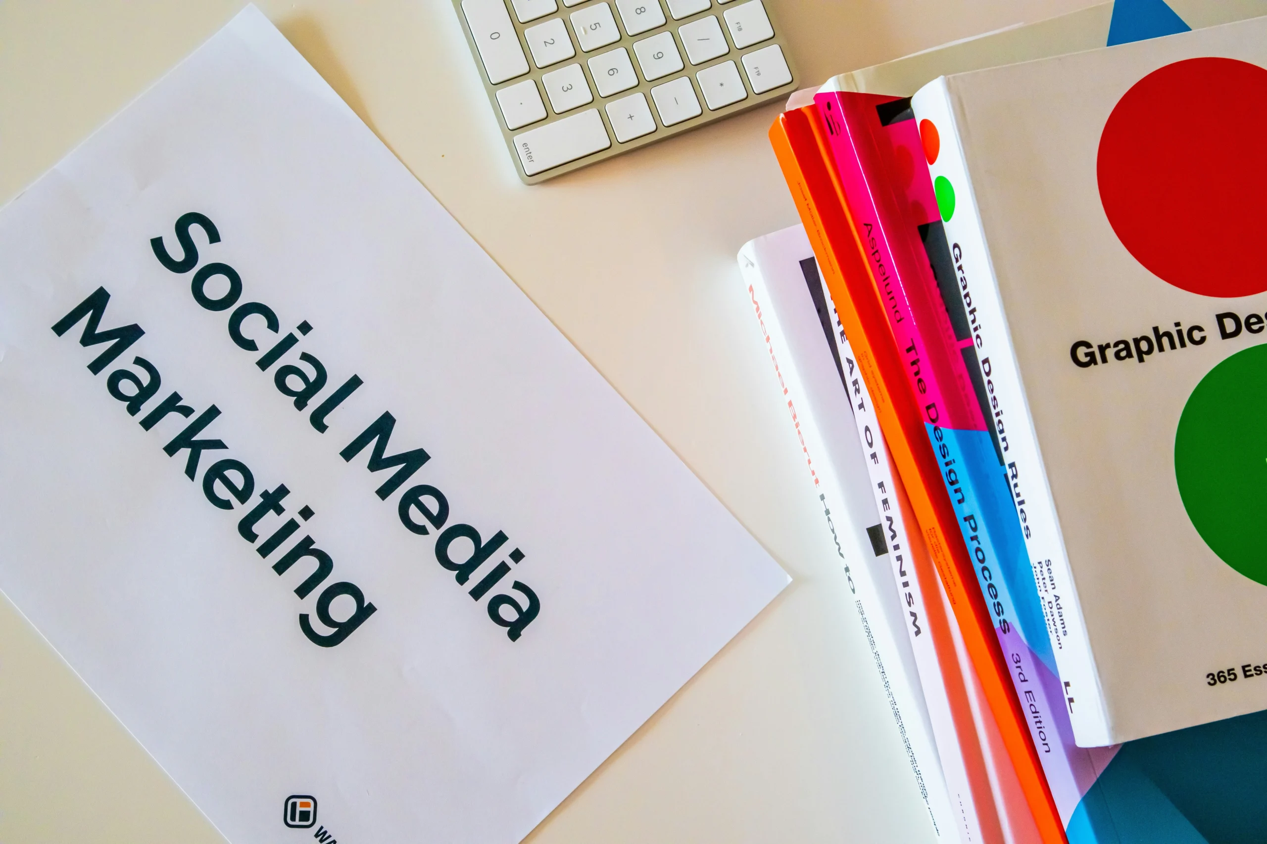 Overhead view of a desk with a printed "Social Media Marketing" document, a white keyboard, and a stack of colorful design books, representing the content planning and strategy work a virtual assistant can manage for small businesses.